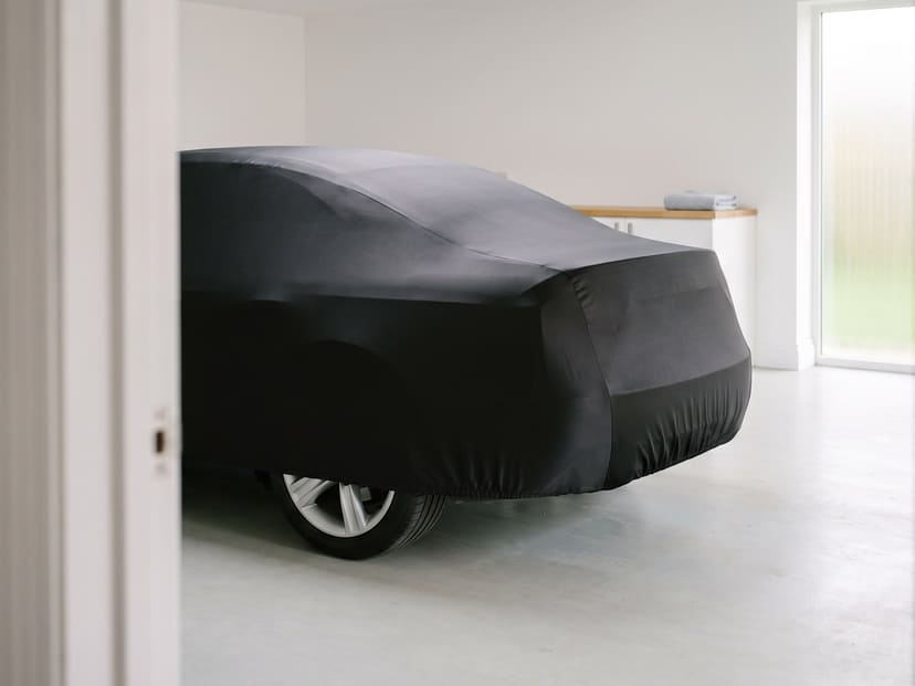 The rear of a sedan under a black stretch cover in a bright white home garage, seen past an open doorway.