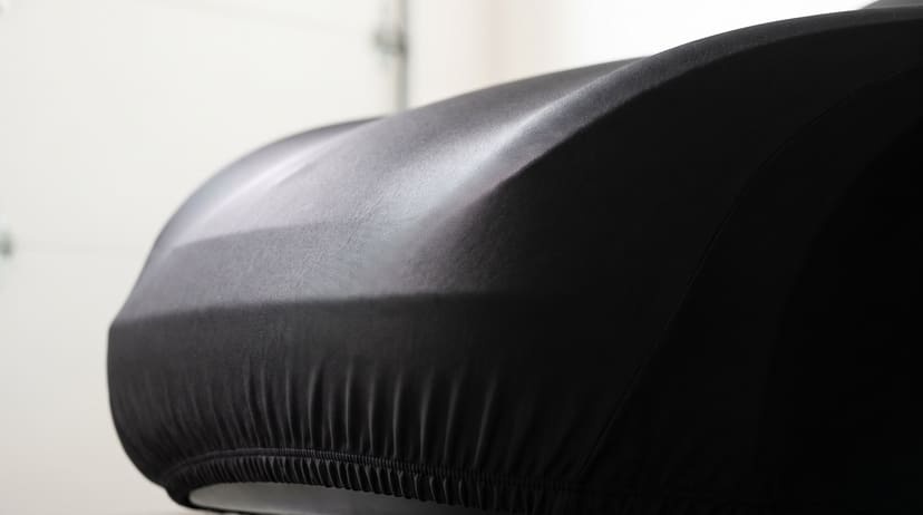 A car under a fitted black satin cover in a bright garage, light sliding along the sheen of the fabric and the elastic hem gathered under the bumper.