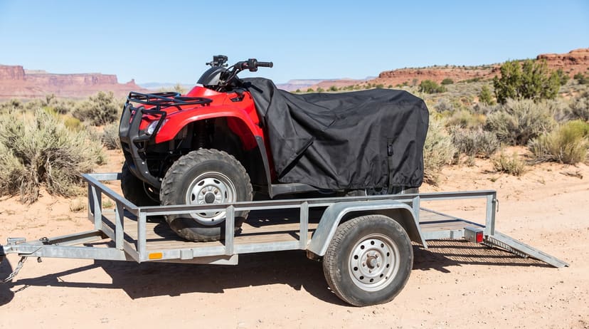 A red ATV on a utility trailer at a desert trailhead, its black cover drawn back off the front over the seat and rear rack.