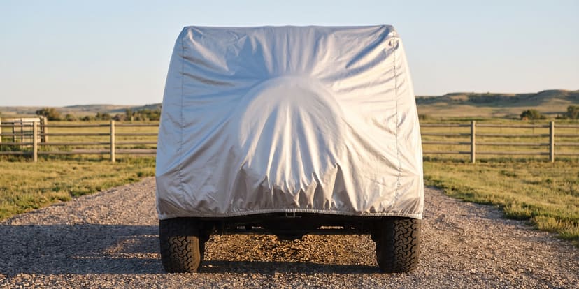 The rear of a big boxy off-road SUV under a silver cover on a gravel ranch drive between split-rail fences, low hills behind in late sun.