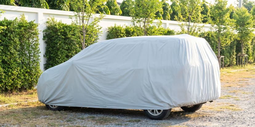 A minivan under a light grey Weatherproof Max cover on a gravel lot beside a white garden wall and hedges