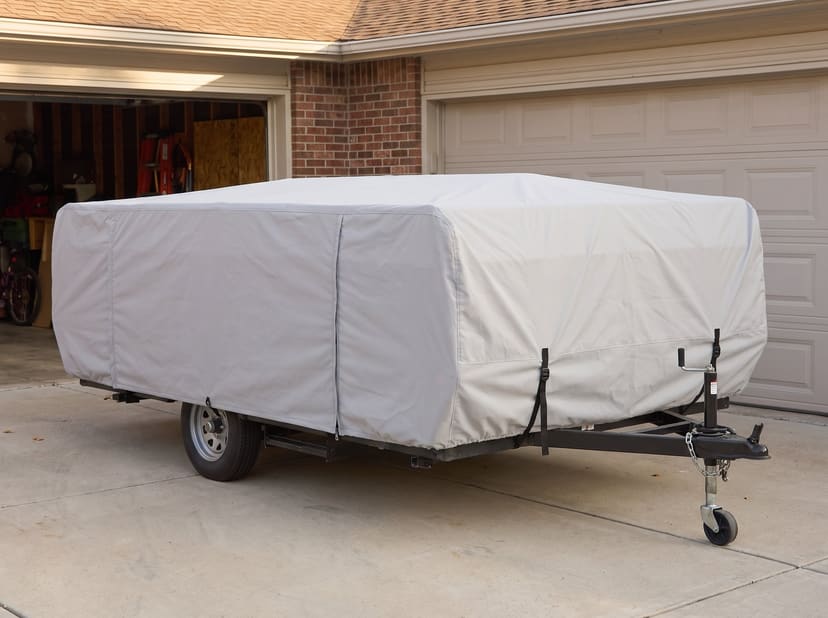 A folded pop-up camping trailer on a driveway in front of a garage, under a fitted light grey cover.