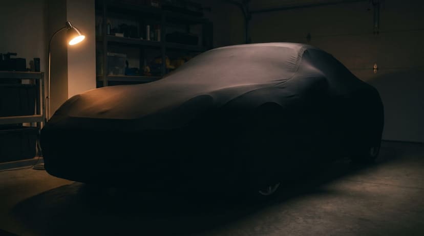A car under a black stretch cover in a dark home garage at night, lit by one floor lamp.