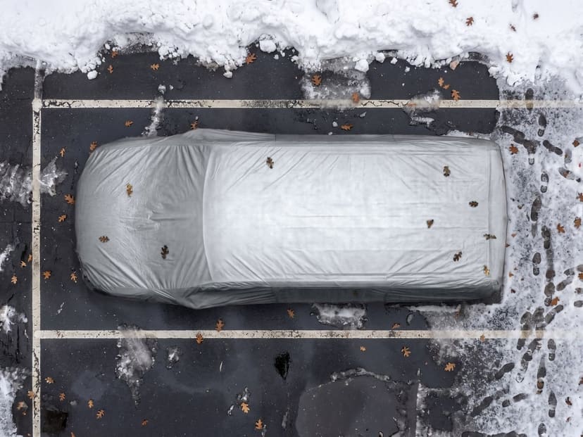 Top-down view of a silver reflective cover fitted over a minivan in a snowy parking space with painted lines and a few fallen leaves.