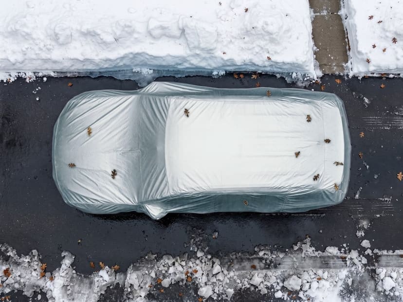 Top-down view of a silver reflective cover fitted over an SUV on dark pavement with snow banks at the edges and a few fallen leaves.
