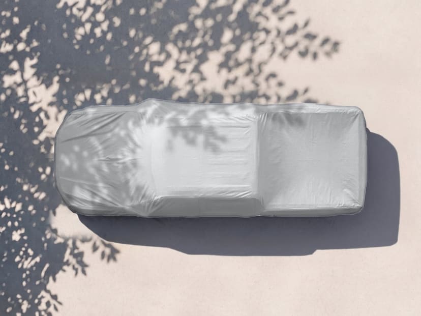 Top-down view of a light grey cover fitted over a pickup truck on pale pavement with tree shade and a long shadow.