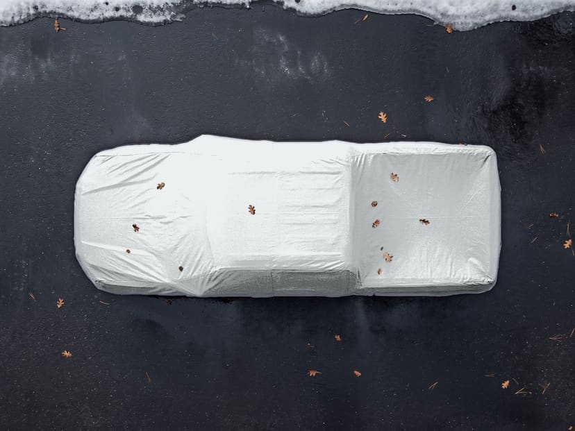 Top-down view of a silver reflective cover fitted over a pickup truck on dark pavement with a little snow at the edge and a few fallen leaves.