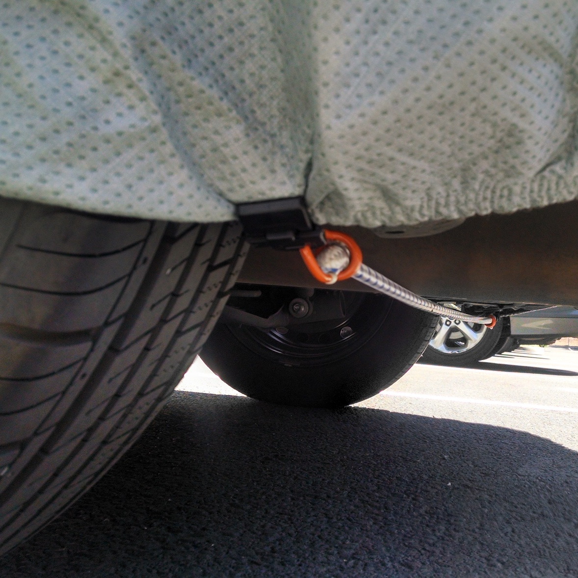 A Gust Strap grip clip on the cover hem, its bungee tensioned under the car, seen from the ground.