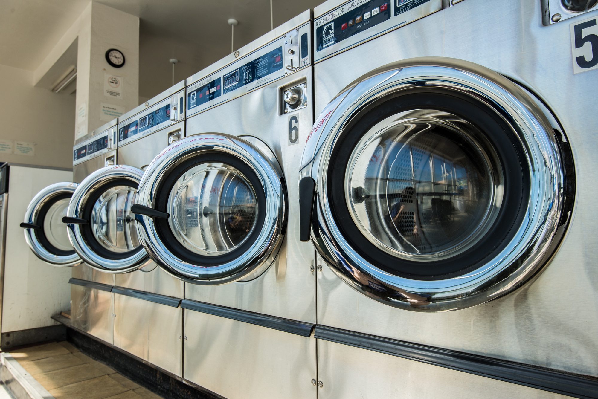 A row of large stainless commercial front-loading washing machines at a laundromat, doors swung open