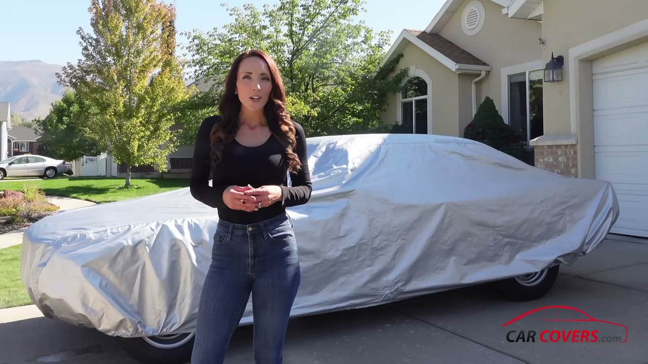 A red classic Mustang fully covered by a silver Platinum Shield cover in a driveway, the presenter at the left